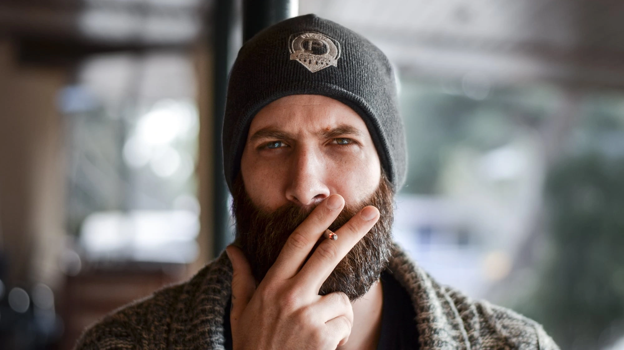 Close-up portrait of a bearded man wearing a knit beanie smoking a lit cigarette.