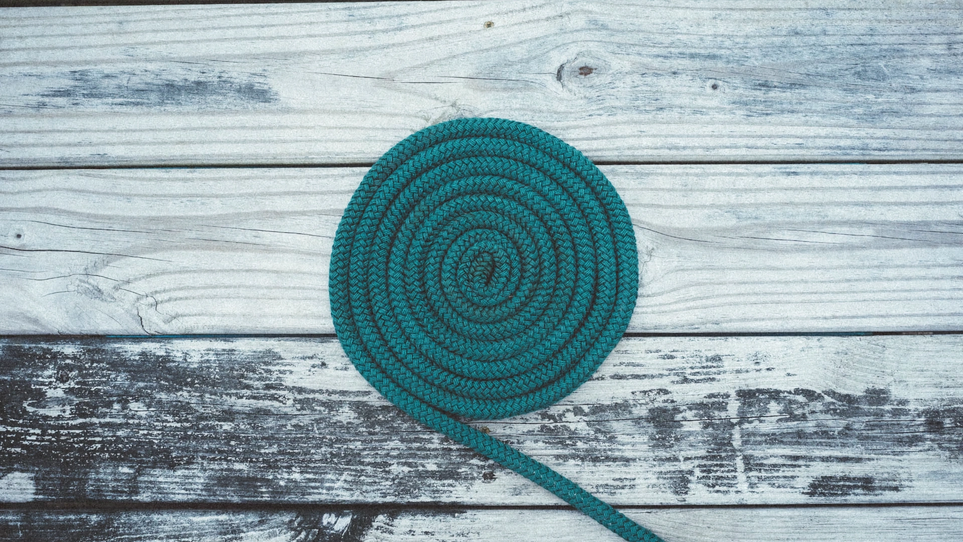 Overhead view of a coiled turquoise rope lying neatly in a flat spiral on weathered grey wooden decking planks.