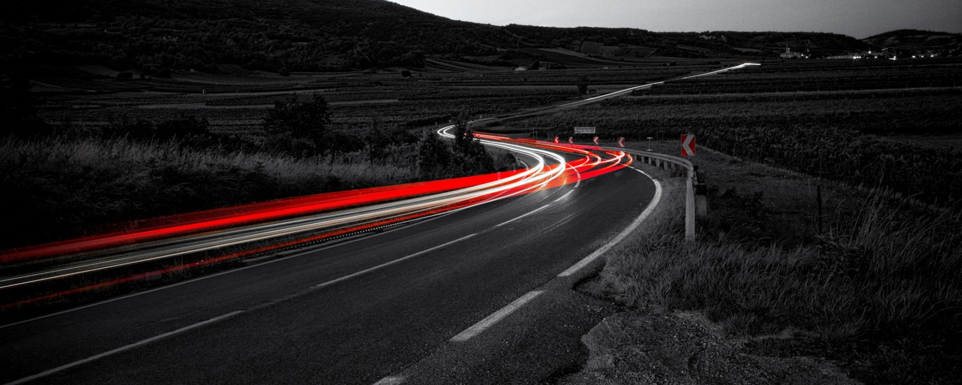 Long exposure photograph at night of a winding road in black and white with vivid red and white car light streaks, illustrating continuous motion and velocity over time
