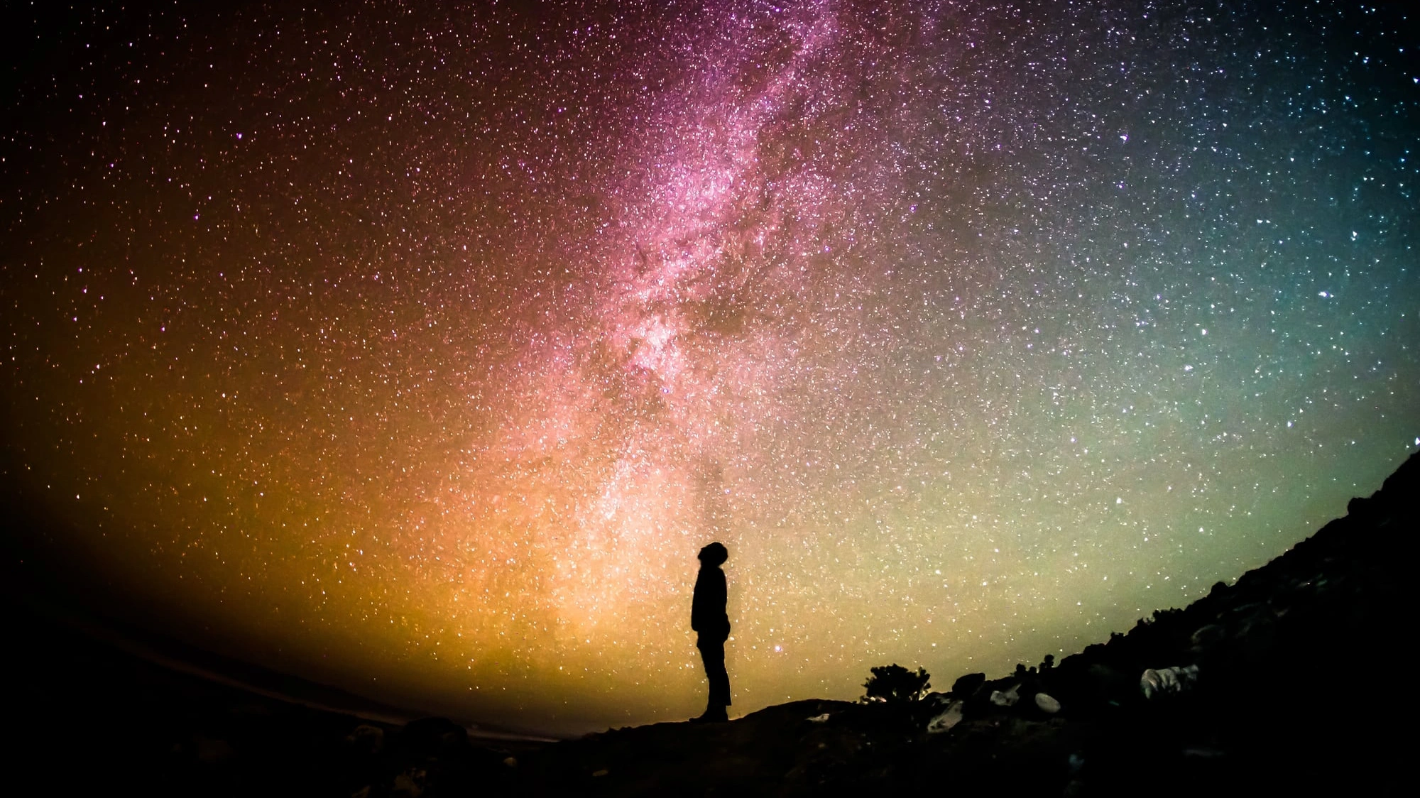 Silhouette of a person standing on a dark ridge gazing up at the vibrant, glowing arch of the Milky Way across a starry night sky.