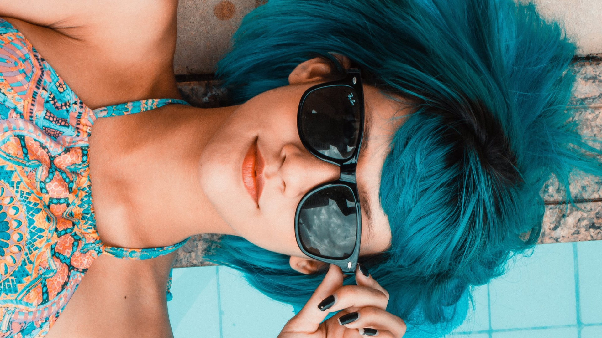 Top-down portrait of a relaxed woman with bright turquoise hair wearing dark sunglasses while lying by the poolside.