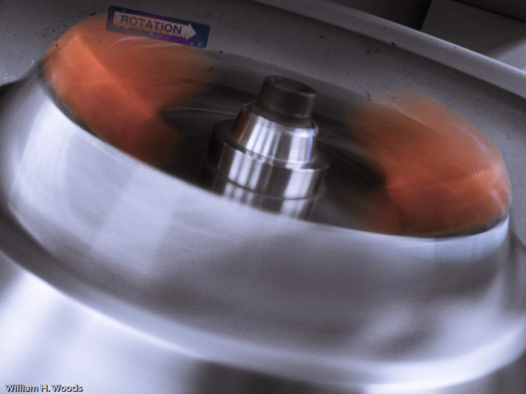 Close-up photograph of a metal centrifuge rotor spinning at high speed, creating motion blur around the central drive spindle.