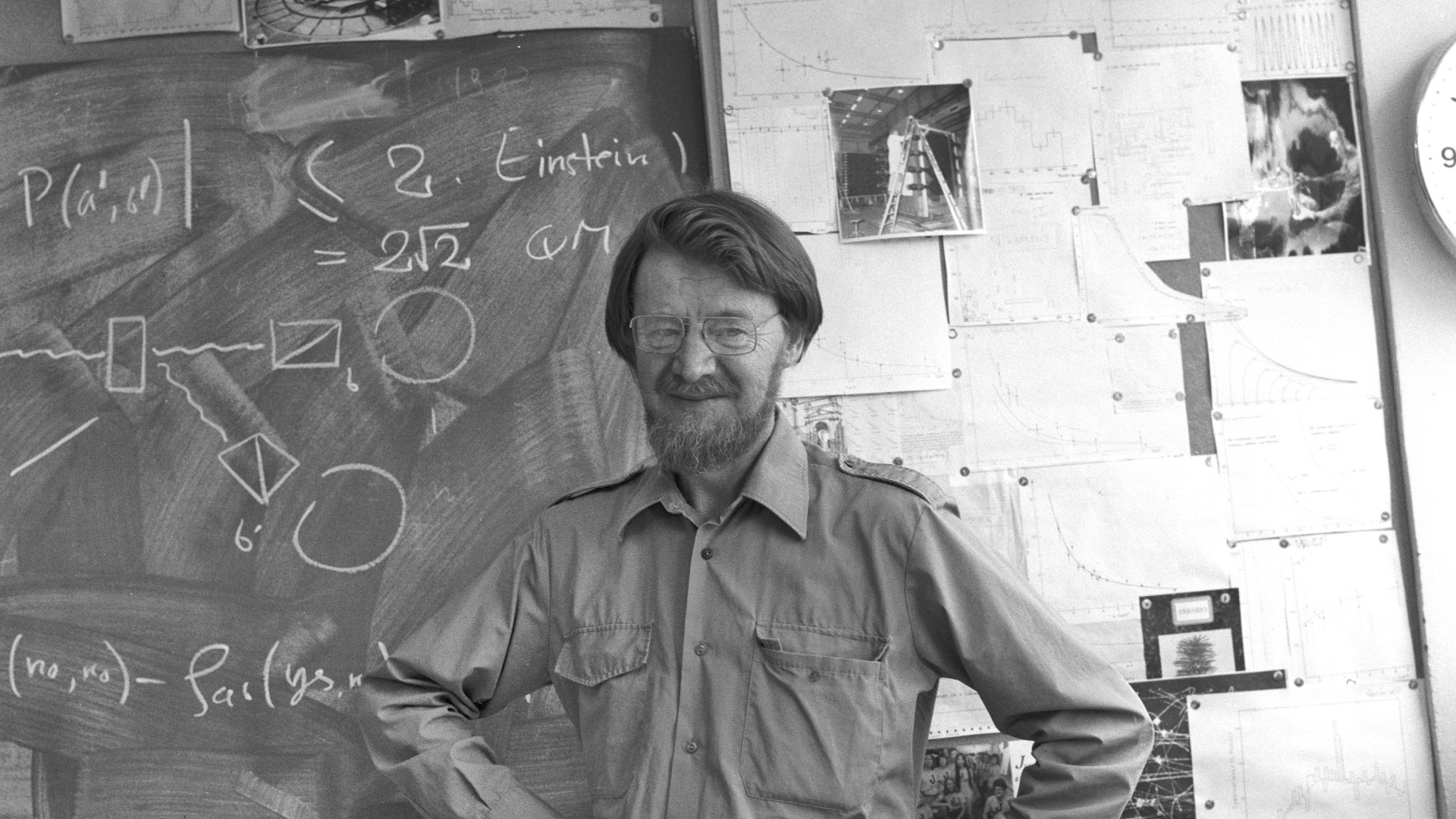 Black and white photograph of theoretical physicist John Stewart Bell smiling with hands on his hips in front of a chalkboard covered in physics equations at CERN.
