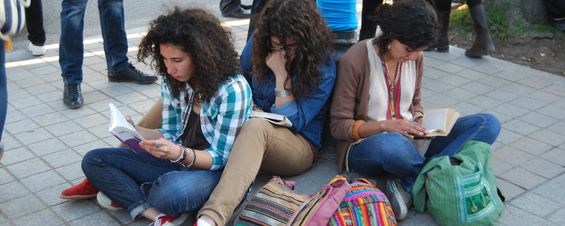 Three young women with dark curly hair sit on the ground of a paved street reading books, with colourful woven bags in front of them.