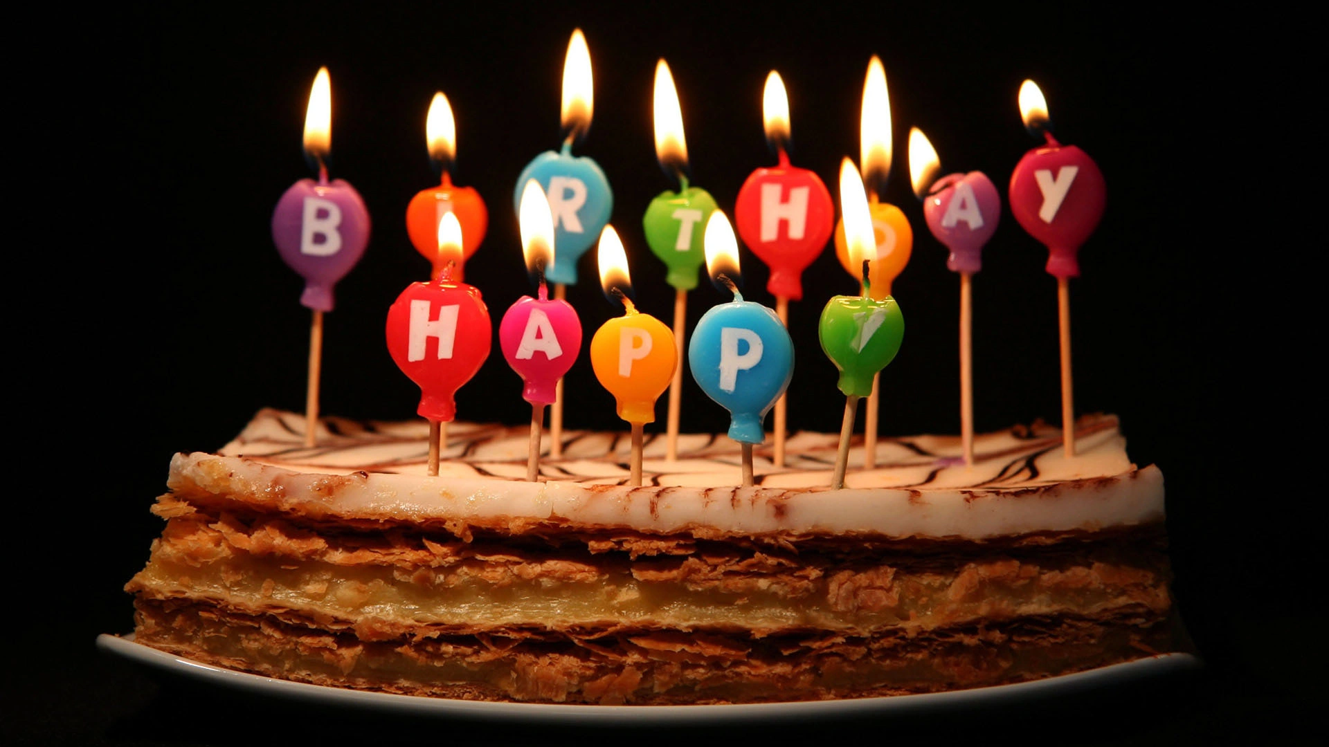 Close-up of a festive mille-feuille birthday cake topped with lit colourful letter candles spelling HAPPY BIRTHDAY against a dark background.