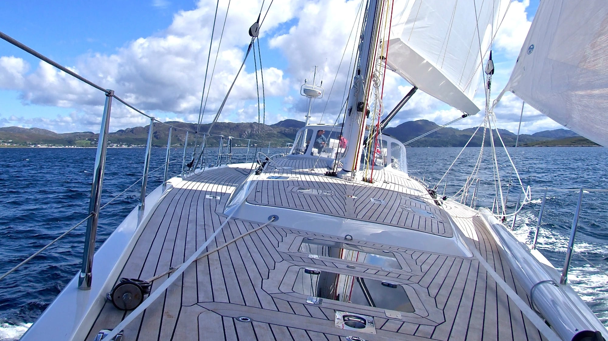 View from the teak deck of a sailing yacht under full white sail on deep blue open waters with rolling green hills in the distance.