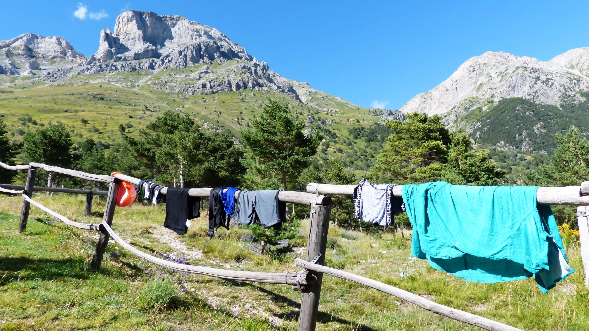 Laundry hanging out to dry on a rustic wooden post-and-rail fence with lush green mountain slopes and rugged rocky peaks in the background.