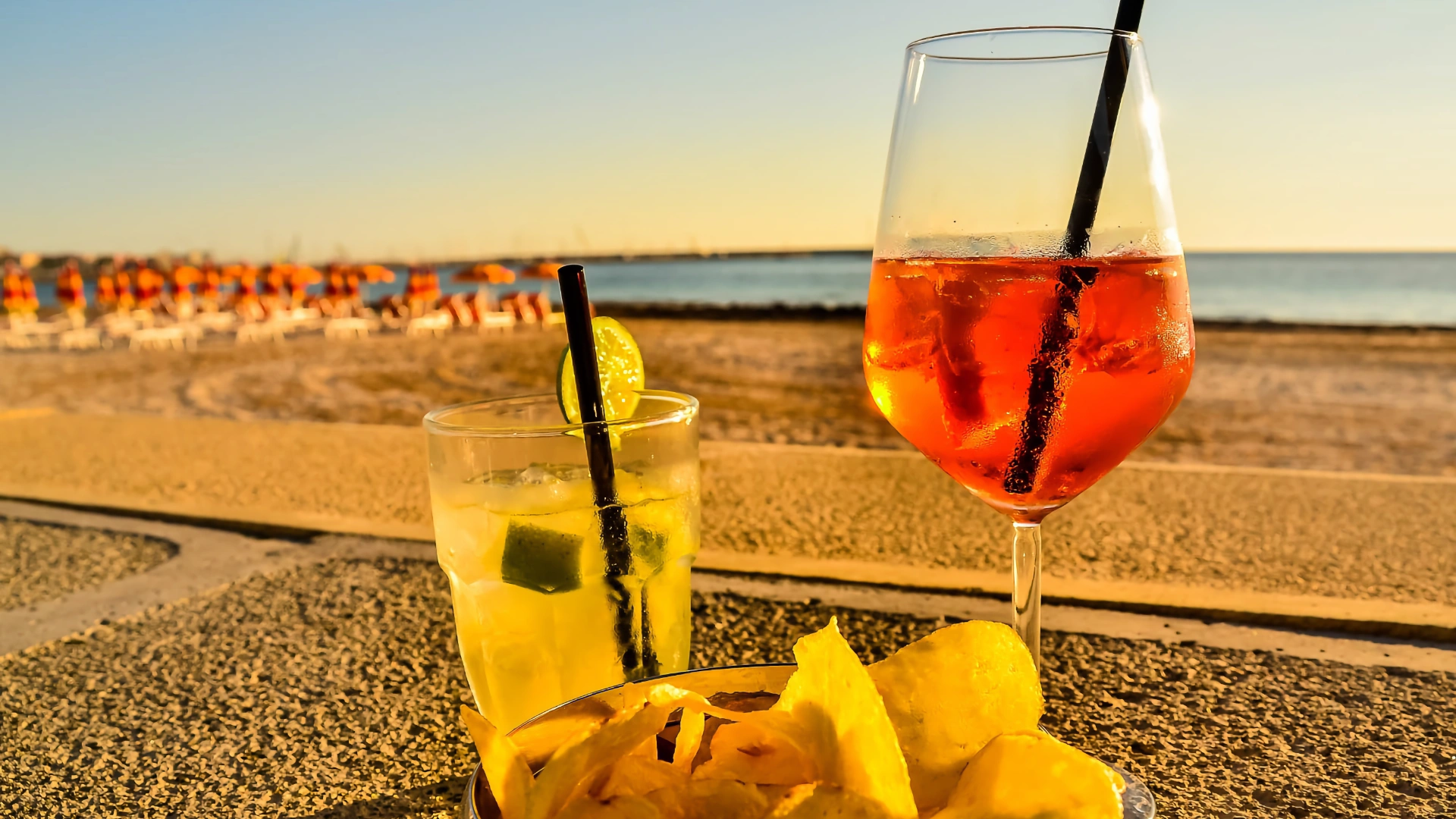 Close-up of a cocktail glass with a black drinking straw, a cold beverage, and crisps on a sunlit beach terrace during sunset.